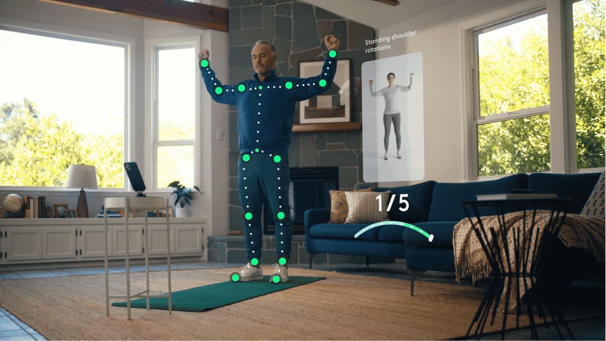 3D pose-estimation skeleton overlaid on a man performing a standing shoulder rotation exercise at home, guided by an on-screen video demonstration